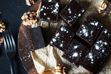 Chocolate brownie next to walnuts and a black fork on a wooden board on a dark background