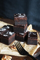 Chocolate brownie next to walnuts and a black fork on a wooden board on a dark background