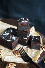 Chocolate brownie next to walnuts and a black fork on a wooden board on a dark background