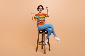Full length portrait of delighted gorgeous person sit chair raise fists attainment isolated on beige color background