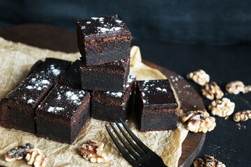 Chocolate brownie next to walnuts and a black fork on a wooden board on a dark background