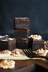 Chocolate brownie next to walnuts and a black fork on a wooden board on a dark background