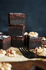 Chocolate brownie next to walnuts and a black fork on a wooden board on a dark background