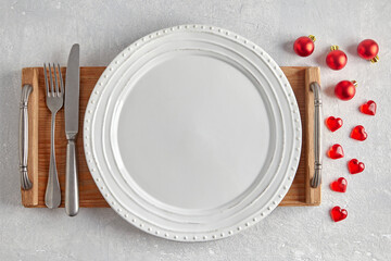Empty white plate with cutlery on a brown wooden tray on a gray concrete table surrounded by red Christmas tree balls and hearts. Template for presenting food in a menu