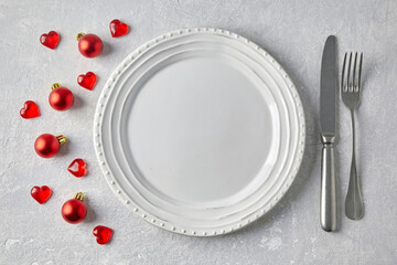Empty white plate with cutlery on a gray concrete table surrounded by red Christmas balls and hearts. Template for displaying food on the menu