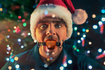Man with mandarin in his mouth as BDSM sex toys on the Christmas tree background. Concept of...