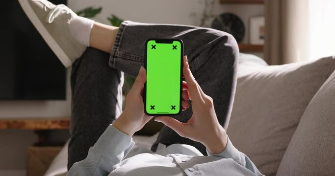 Woman explores endless potential of smartphone green screen chromakey resting on sofa, vertical orientation. Advertising app. Tap in center to watching videos, relaxation, entertainment, digital age