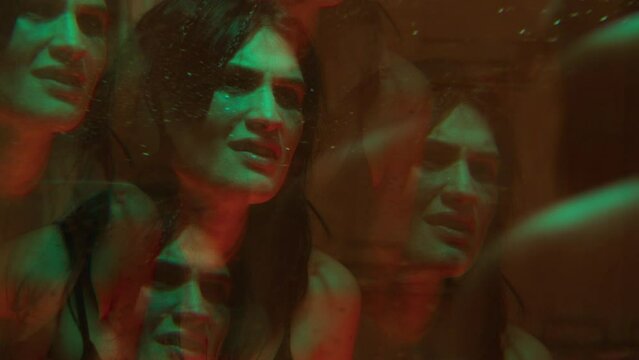 Anxious woman looking at her reflection in wet mirror, rubbing her face and feeling scared in a bathroom with multi-colored neon light. Kaleidoscope effect, mental problem concept