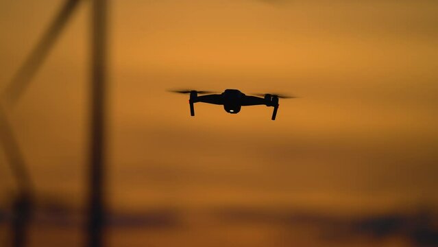 Drone flying against Silhouette Wind turbine rotating by wind, quadrocopter uses for study advantages and drawbacks of electric station at incredible multi colored cloudy sunset at evening