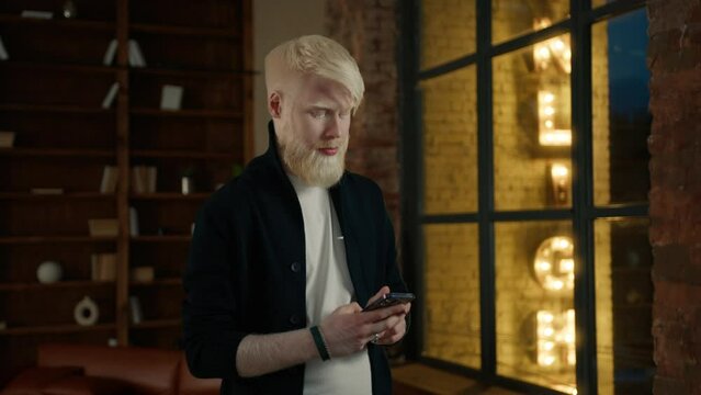 Focused albino businessman typing message on smartphone in loft office with night city lights seen in large window on motion background. Male hipster mobile phone. Casual freelancer working at night