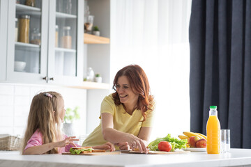 A friendly family from mother and daughter manage in the kitchen in a joyful mood. A young woman mom and little girl smile and discusses various delicious culinary recipes to make sandwiches. The - obrazy, fototapety, plakaty
