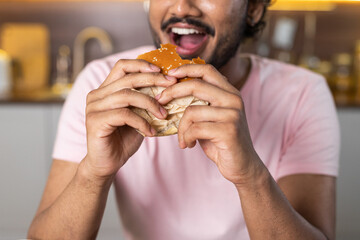 A young african or american man in the kitchen with open mouth holds a hamburger in hands and eats...