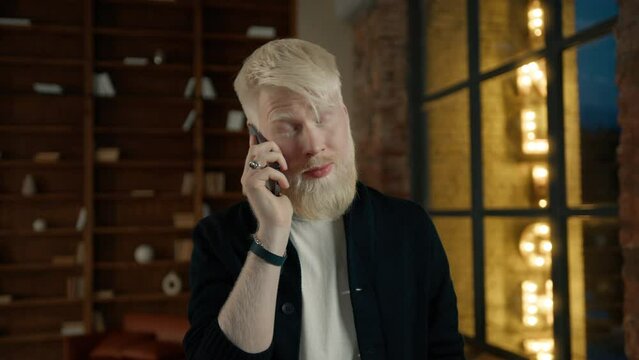 Annoyed albino businessman arguing in smartphone loudspeaker. Angry businessman talking on mobile phone in loft office. Casual employee working late at night in loft. Casual guy feeling stress at work