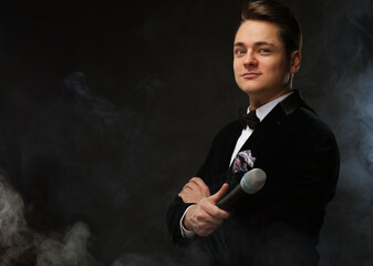 Stylish man in a tuxedo holding a microphone, posing against a dark background with smoke