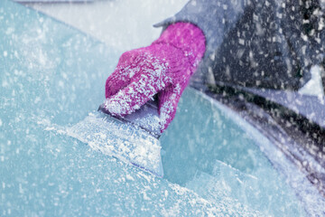 Winter Driving - Close-up of a woman's gloved hand using an ice scraper to remove ice from the frozen windscreen of her car before driving.