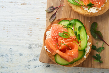 Open smoked salmon sandwiches with cream cheese, cucumber, sesame seeds, microgreens, spinach, and peas leaves on light old wooden background. Healthy breakfast food. Delicious snack. Top view.