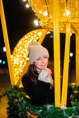 Beautiful woman in warm clothes, a hat and a coat in winter against the background of Christmas street lights. Celebrates the New Year on the street against the backdrop of street decor. bokeh