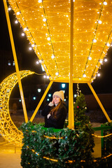 Beautiful woman in warm clothes, a hat and a coat in winter against the background of Christmas street lights. Celebrates the New Year on the street against the backdrop of street decor. bokeh