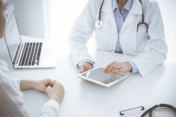 Fototapeta premium Doctor and patient sitting opposite each other at the desk in clinic. The focus is on female physician's hands pointing into tablet computer touchpad, close up. Medicine concept