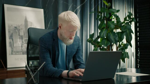 Portrait of albino businessman working on laptop at the office table. Male executive reading email on computer screen. Serious manager feeling surprised. Shocked worker using pc in the office 
