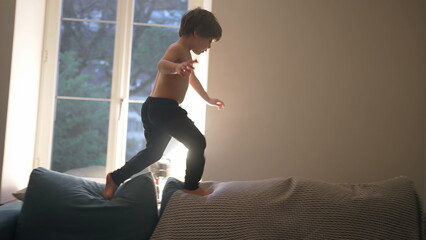 Child having fun at home by himself jumping and running on top of couch sofa, energetic carefree kid