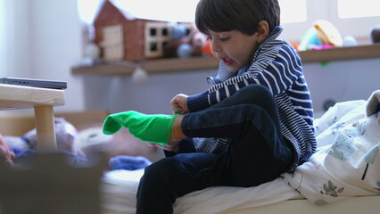 Child putting on sock seated by bedside. Small boy dressing himself. Kid getting ready to start the...