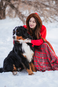 woman in winter in an ancient red coat and plaid skirt in winter in the snow with a dog of the Bernese Mountain Dog breed. Fairytale image of Little Red Riding Hood with a big dog.