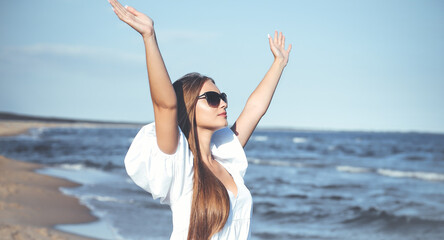 Happy blonde woman is on the ocean beach in a white dress and sunglasses, raising hands