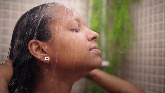 Young black woman taking relaxing shower at home. African American female wetting her hair and head with water enjoying self-care in bathroom. Concept of daily domestic personal hygiene in morning.