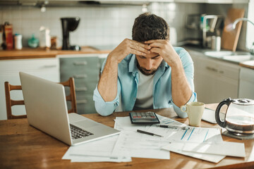 Stressed young man managing debt in kitchen - obrazy, fototapety, plakaty