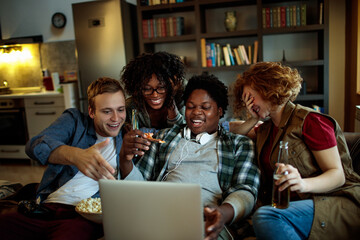 Multicultural group of young friends having snacks and watching something on laptop in living room
