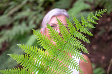 Defocused smiling woman in trekking day in the forest partially hidden by a fern leaf. Smiling retired lady looking at camera enjoying nature and outdoors