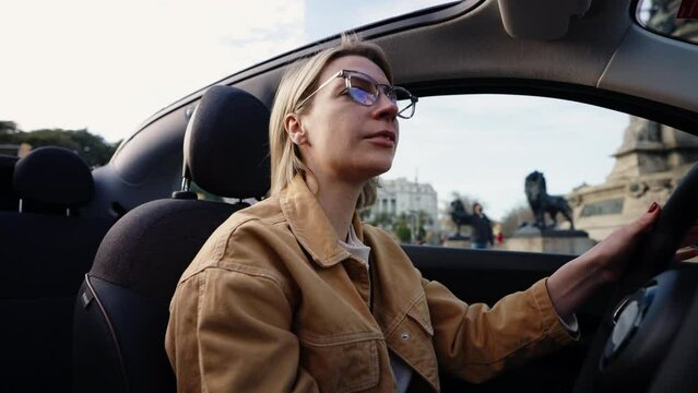 Pensive woman in casual wear driving her eco-friendly car, contemplating the ease of navigating through city's landmarks on a clear day. Focused female tourist ride on Barcelona streets on rental auto