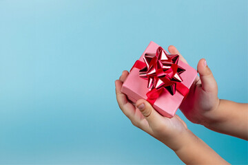Mãos de uma criança segurando um presente com embalagem cor-de-rosa em um fundo azul claro. - obrazy, fototapety, plakaty