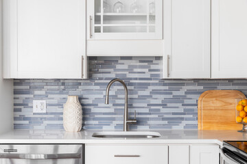 A kitchen faucet detail with a blue glass tile backsplash, white cabinets, decorations on the...