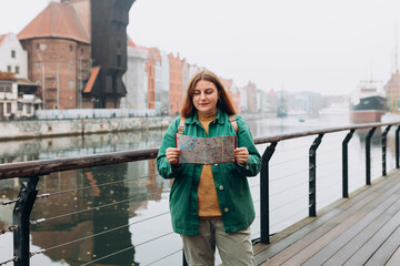 Attractive young female tourist is exploring new city. Redhead girl with backpack holding a paper map on city street in Gdansk. Traveling Europe in autumn. Famous Zuraw crane, Motlawa river