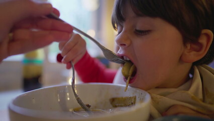 Mother feeding child spaghetti pasta food on bowl covered with napkin on collar. Little boy enjoys Italian food