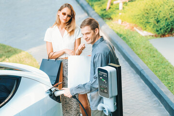 Young couple travel with EV electric car charging in green sustainable city outdoor garden in...