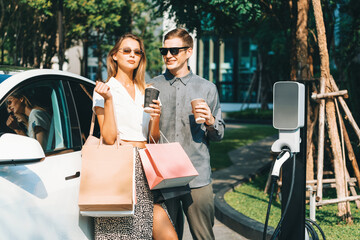Young couple travel with EV electric car charging in green sustainable city outdoor garden in...