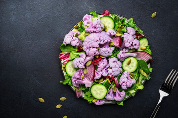 Obraz na płótnie Canvas Healthy fresh vegan salad from purple cauliflower, cucumbers, red onion, radicchio, spinach with pumpkin seeds, dark table background, top view