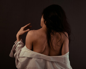 Back view of sexy young long hair brunette woman bares her back and shoulder taking off the white men shirt on studio background. Sensual