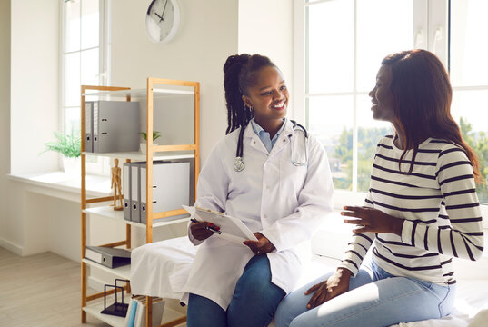 Happy Smiling Woman Patient Talking With Friendly African American Female Doctor Therapist Holding Report File With Appointment Sitting On The Couch In Office During Medical Examination In Clinic.