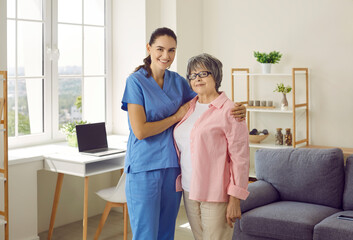 Young smiling brunette caregiver or nurse supporting and hugging senior woman patient at home. They...