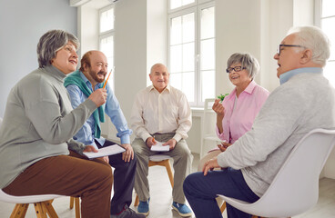 Mental health of elderly. Cheerful elderly people are talking at psychological therapy in nursing home. Smiling mature men and women sitting in circle on chairs, talking and making notes in notebook. - obrazy, fototapety, plakaty