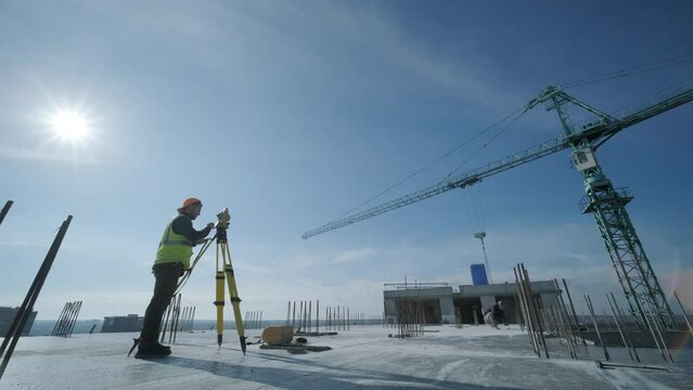 Geodetic works. Surveyor engineer in protective wear and red helmet using geodetic equipment at construction site. Professional equipment.