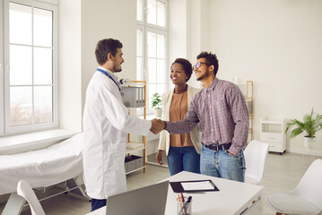 Smiling young multiracial couple handshake greeting with male doctor at consultation in clinic. Happy ethnic man and woman spouses shake hand make deal with GP in hospital. Healthcare concept. - obrazy, fototapety, plakaty