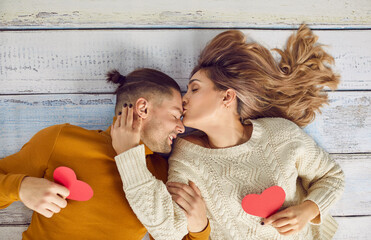 Romantic photo session of a couple in love on Valentine's Day. Young woman kisses her man on the forehead while lying with heart shaped cards on a rustic white wooden floor. Top view, shot from above