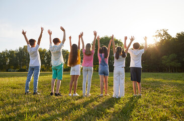 Group of kids friends raising hands up on green grass in the park standing back in a line at sunset. Children having fun together outdoors on a sunny summer day in casual clothes in nature.