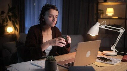 Tired freelancer having remote work on laptop checking email and drinking hot coffee or tea at night Busy young woman student having online distance education on computer at home workplace