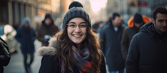 A happy woman is pictured in a black winter coat as she walks down a city street embodying the hipster lifestyle and enjoying her autumn vacation surrounded by people of different background - obrazy, fototapety, plakaty